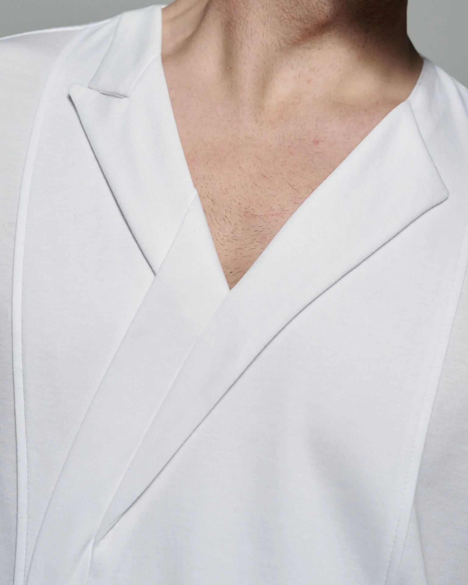 This image features a close-up of the W. Rubens 73, an Asymmetric V-neck T-shirt with Lapels & Inserts, elegantly showcasing its unique design elements. The T-shirt, crafted in England, highlights the brand's commitment to redefining conventional style through innovative design. The V-neckline is framed by asymmetric lapels, where a peak lapel graces the left side, offering a structured and refined appearance, while the right side exhibits a collarless design, creating an avant-garde and contemporary look. The image also reveals the artful folded insert on the front body panel, a distinctive feature that enhances the T-shirt's aesthetic and provides a sense of dimension and texture. The contrast between the soft, smooth white cotton fabric and the precise grey top stitching adds a touch of finesse, elevating the overall design. This stitching not only enhances the visual appeal but also ensures the garment's durability and longevity. The slim block fit, as seen in the image, is designed to contour the body without being restrictive, offering a comfortable and flattering silhouette. The fit is further refined by the French side seams, which maintain the T-shirt's polished appearance and ensure that it holds its shape over time. The choice of fabric and craftsmanship ensures that this T-shirt feels luxurious against the skin, offering both comfort and style.